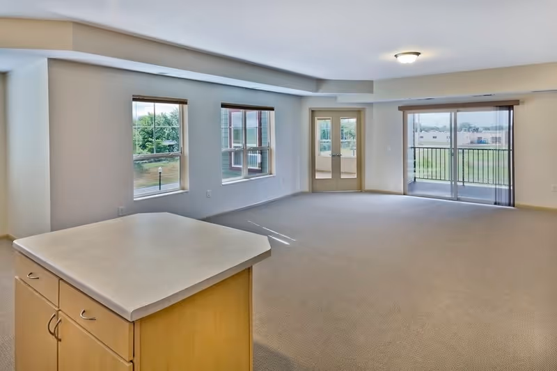 Spacious empty living area with beige carpet and light-colored walls, featuring three windows, a glass door leading to a balcony, and a kitchen island with cabinets in the foreground.