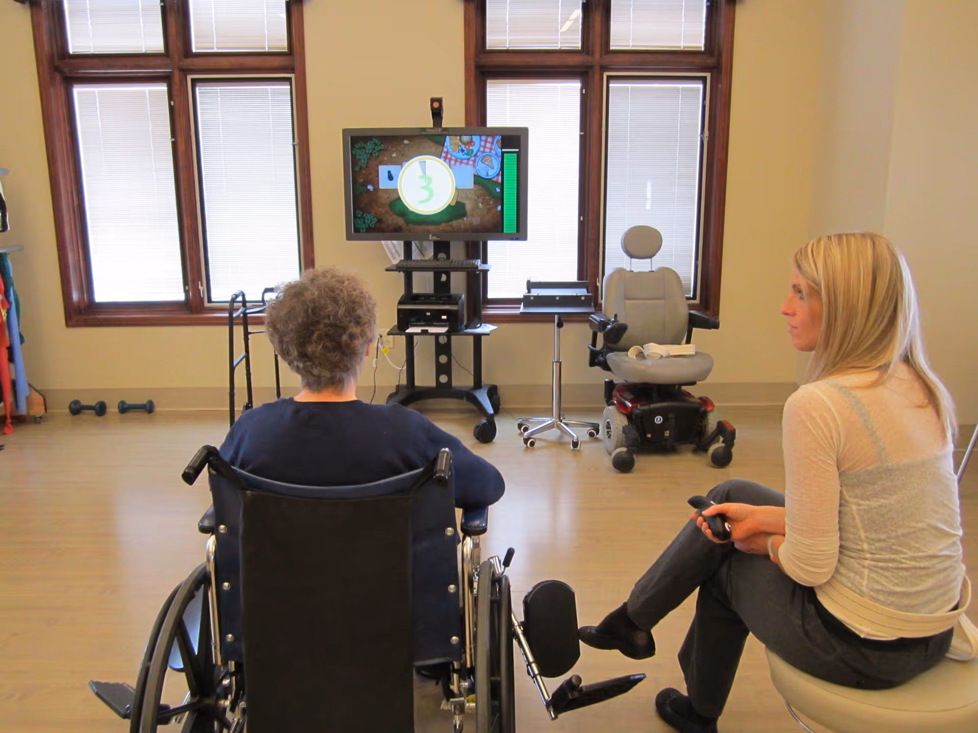 An elderly person in a wheelchair and a younger woman sitting in a room with large windows. They are facing a television screen mounted on a stand, which displays a game or interactive activity. The room has light-colored walls and wooden flooring, with some exercise equipment and a motorized wheelchair visible in the background.