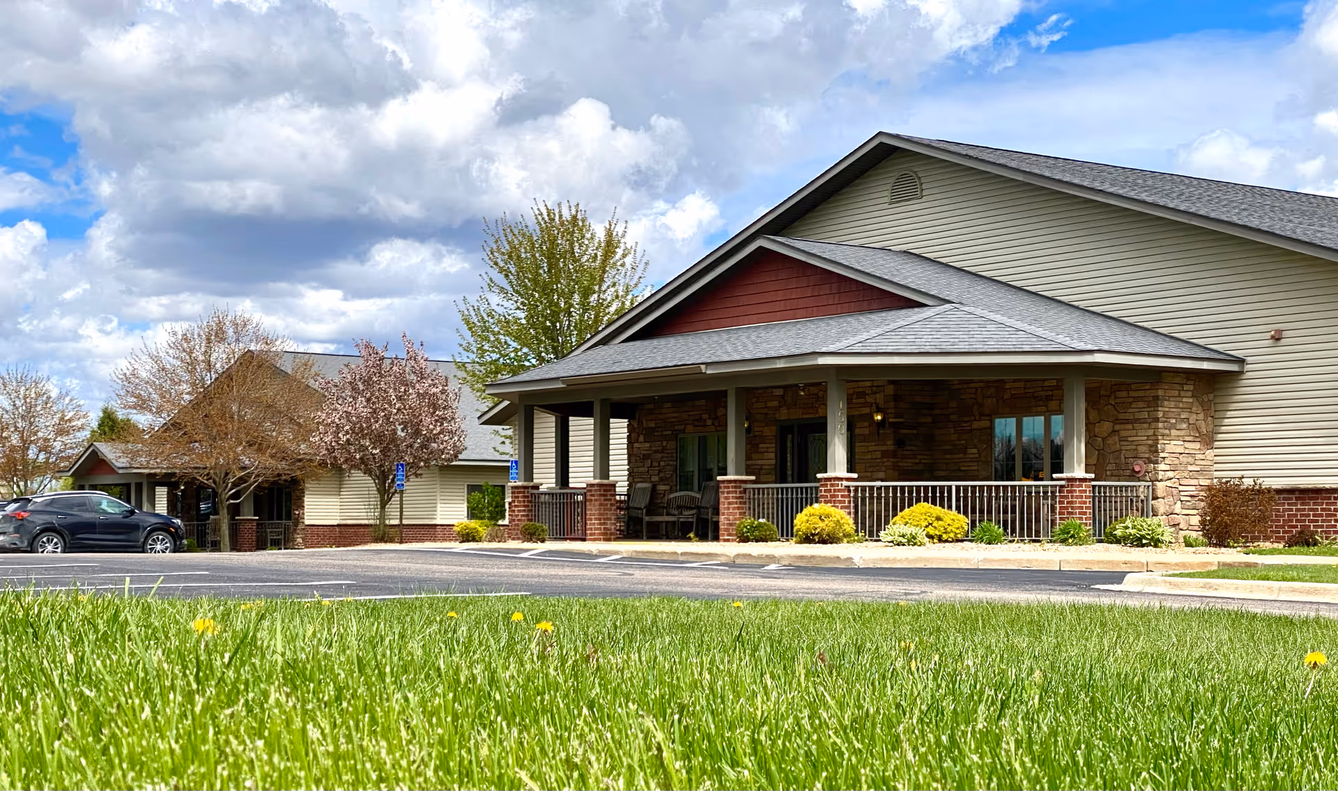 Front exterior of a single-story senior living cottage with stone and siding facade, covered porch, and lawn in the foreground.