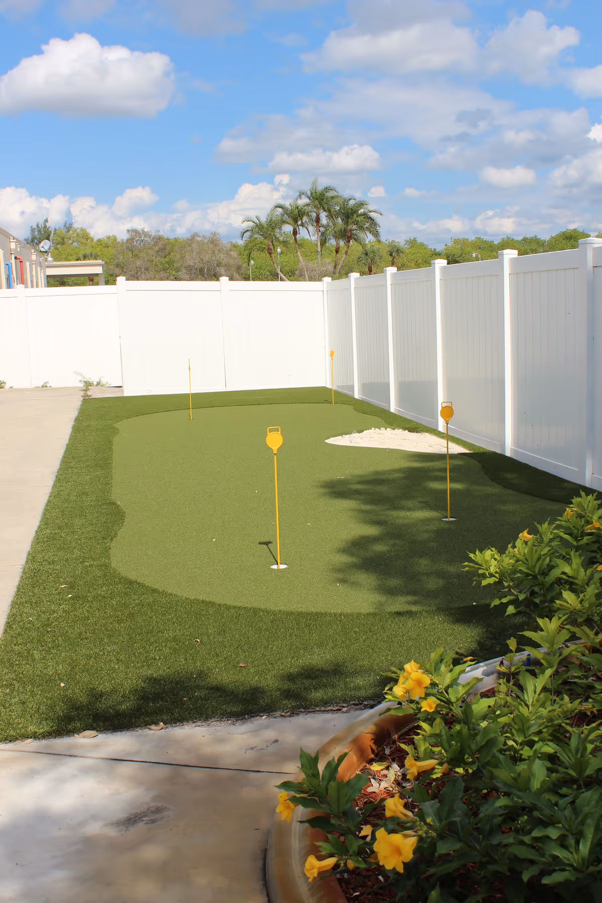 Outdoor putting green area with artificial turf, several yellow flagsticks marking holes, surrounded by a white privacy fence. There are palm trees and other greenery in the background under a partly cloudy blue sky. Yellow flowers and green plants are visible in the foreground near a curved concrete pathway.