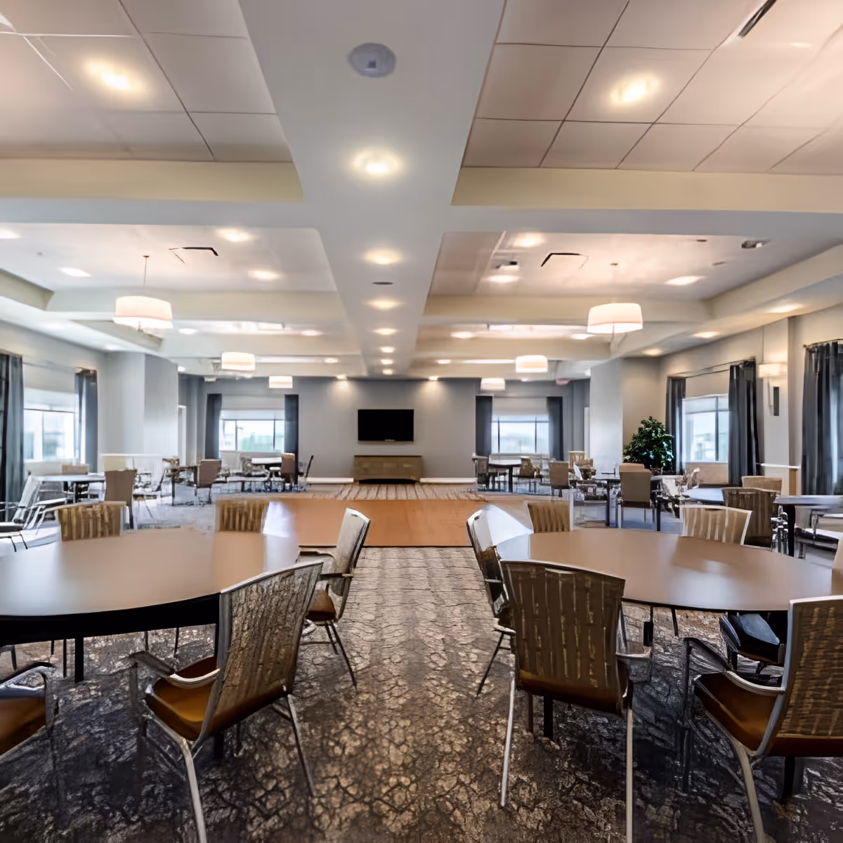 A spacious and well-lit common area with multiple round tables and chairs arranged throughout the room. The room features large windows with curtains, modern ceiling lights, and a flat-screen TV mounted on the far wall above a console table. The carpet has a textured pattern, and there is a small stage or raised platform in the center background.