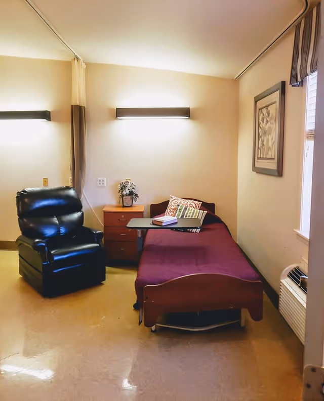 A small bedroom in a senior care facility featuring a single bed with a maroon bedspread and decorative pillows, a black recliner chair, a wooden nightstand with a flower arrangement, a wall-mounted light above the bed, a window with striped curtains, and an air conditioning unit below the window.