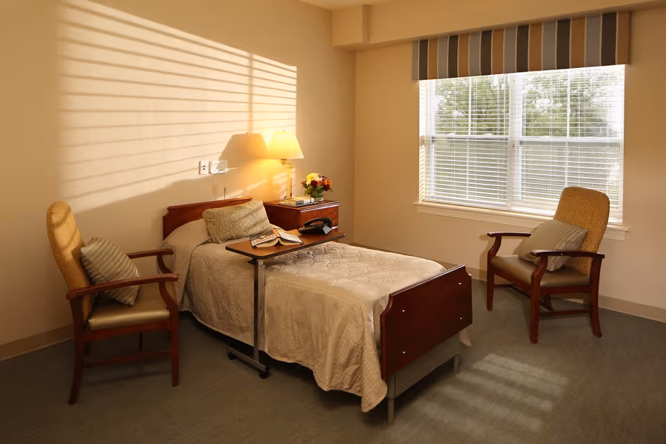 Well-lit senior living bedroom with a single bed, overbed table, two armchairs, nightstand and a window with blinds.