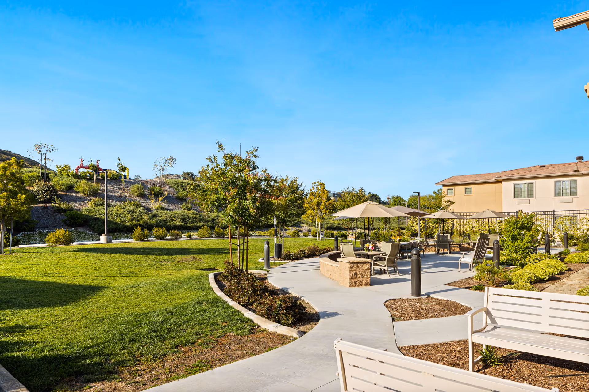 Outdoor patio area at Ivy Park At Murrieta with benches, tables, chairs, and umbrellas surrounded by landscaped greenery and a clear blue sky.
