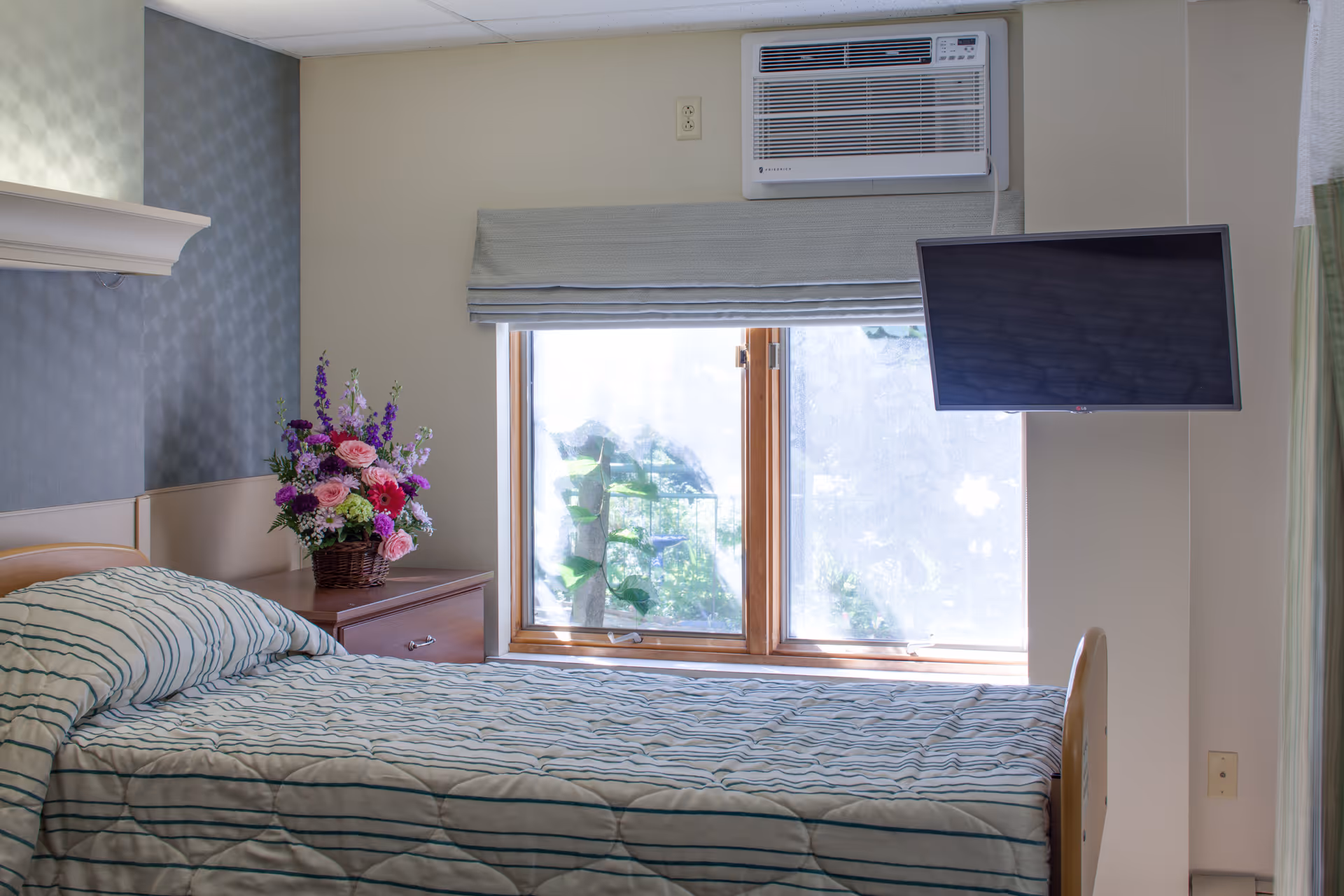 A single bed with a striped bedspread in a room featuring a window with a light blue roman shade, an air conditioning unit above the window, a wall-mounted flat screen TV, and a wooden nightstand with a basket of colorful flowers.