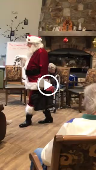 A person dressed as Santa Claus walking in a room with wooden floors and chairs. The room has a stone fireplace decorated with holiday ornaments and a whiteboard in the background. Several seated people are partially visible watching Santa.