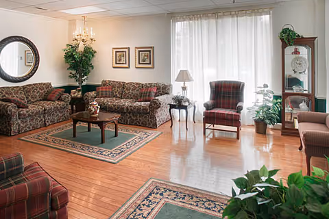 A cozy living room with wooden floors, two floral patterned sofas, a plaid armchair, a wooden coffee table on a green area rug, side tables with lamps, framed artwork on the wall, a round mirror, and several potted plants near large windows with sheer white curtains.