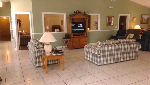 A spacious living room with green walls and white tiled floor featuring two plaid sofas facing a wooden entertainment center with a TV. There is a wooden side table with a lamp and books between the sofas. The room has several framed pictures on the walls and recliner chairs in the background.
