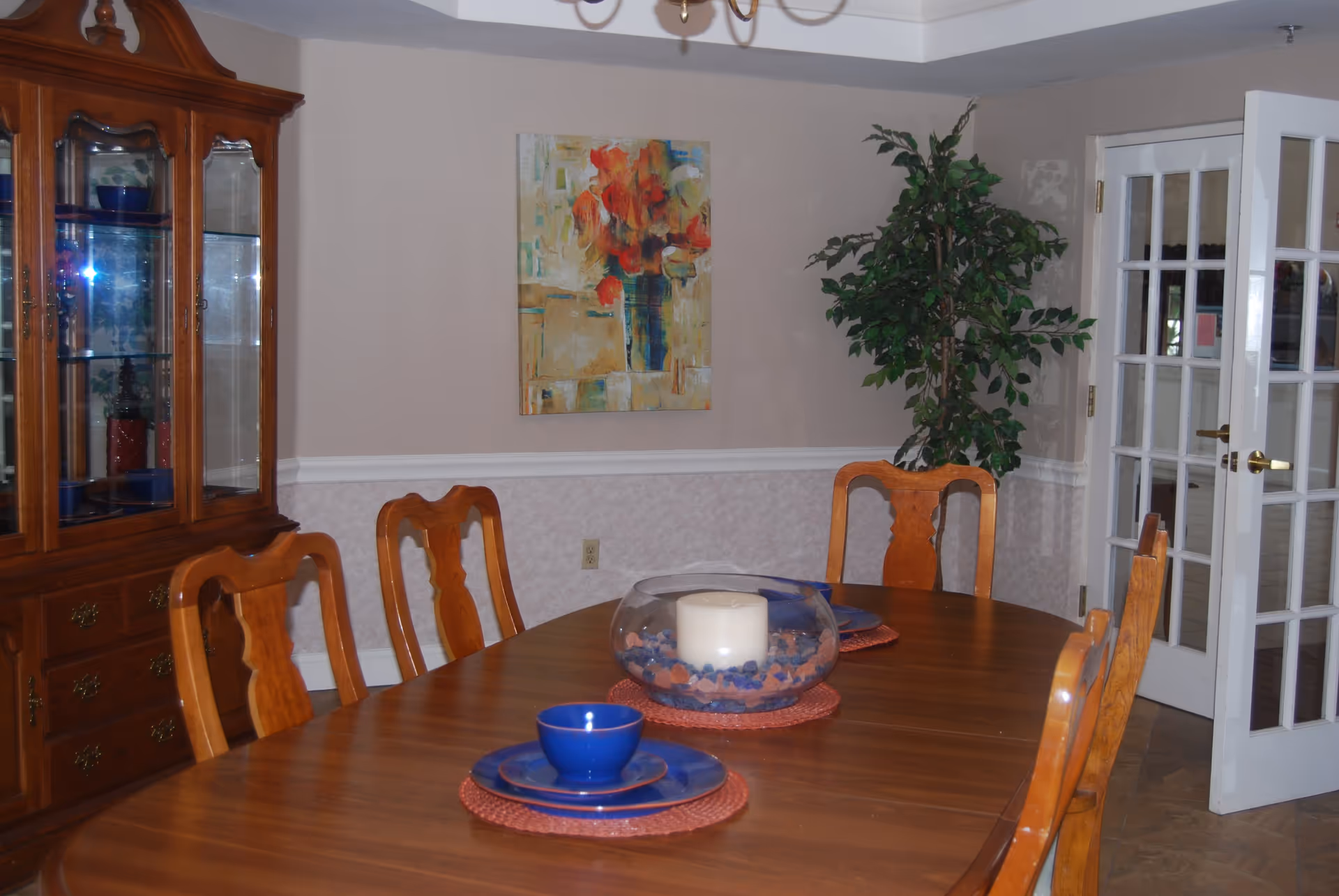 A dining room with a wooden table set with blue dishes and a centerpiece consisting of a large white candle inside a glass bowl filled with decorative stones. The room features wooden chairs, a wooden china cabinet with glass doors, a potted plant in the corner, a colorful abstract painting on the wall, and white French doors.