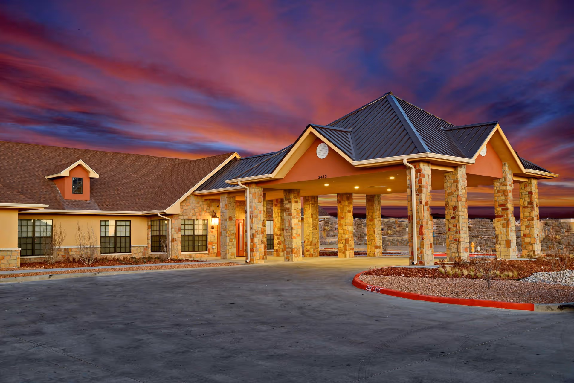 Exterior view of The Suites Rio Vista facility at sunset, showing a building with a covered entrance supported by stone pillars and a driveway in front.