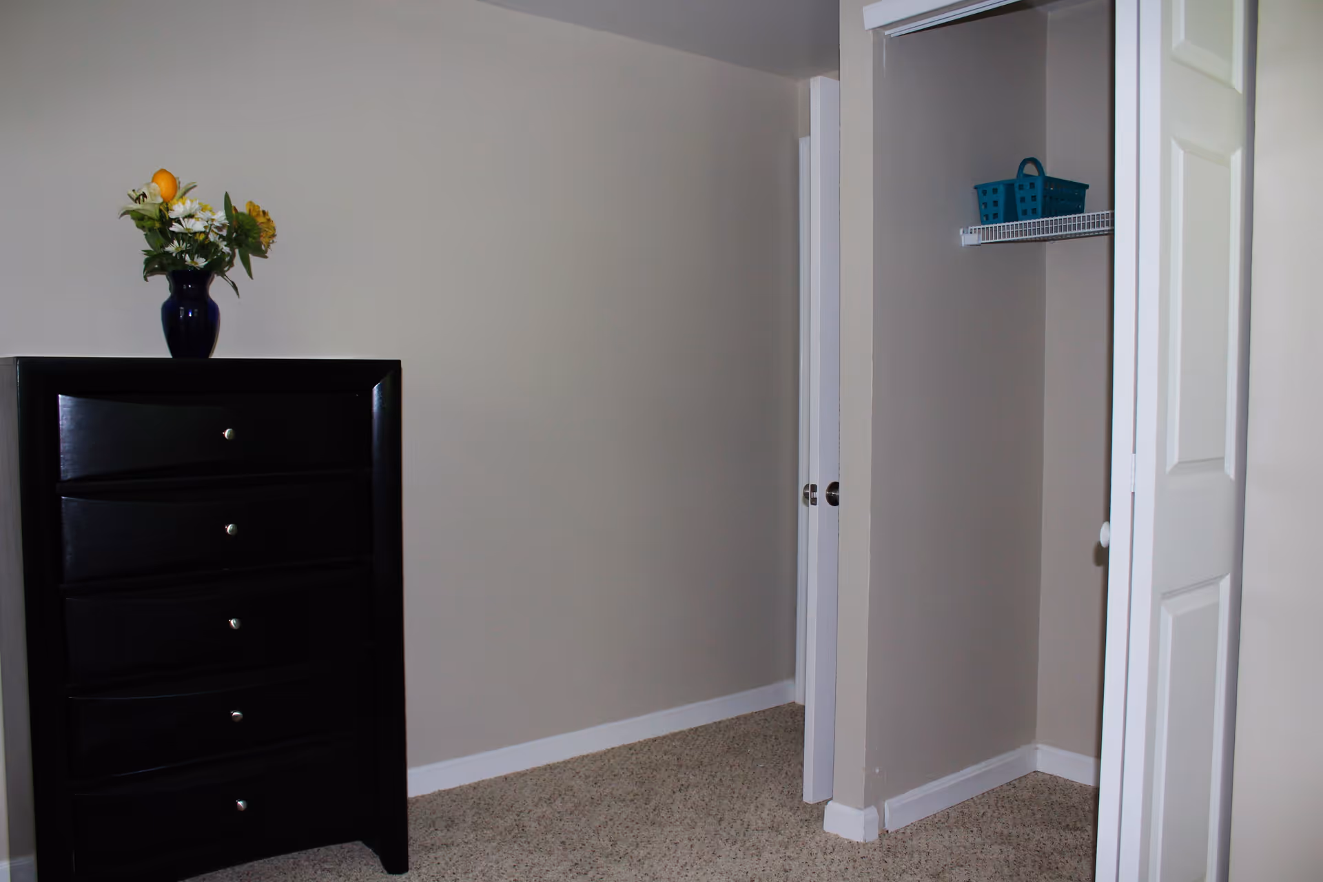 A corner of a bedroom with beige walls and carpeted floor. There is a black chest of drawers with five drawers on the left side, topped with a blue vase containing a bouquet of flowers. On the right side, there is an open closet with a white shelf holding a blue basket.