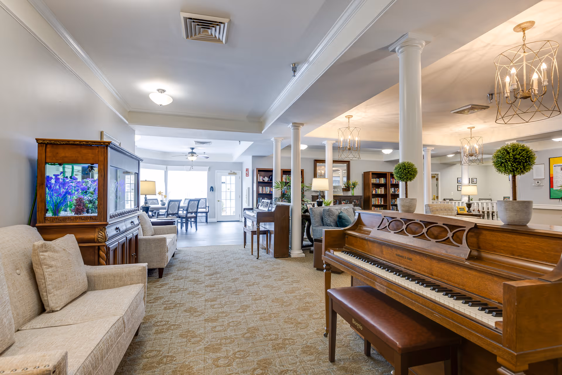 Bright senior living common area with a wooden piano, sofas, an aquarium, white columns, and a dining area in the background.
