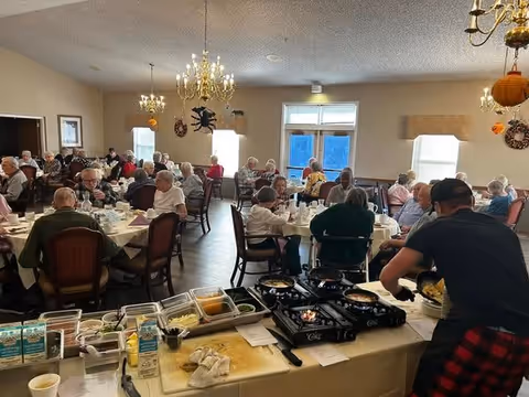 A dining room filled with elderly people sitting at round tables covered with white tablecloths, eating and socializing. A man is cooking food on portable stoves at a serving station in the foreground. The room is decorated with chandeliers, wall wreaths, and has windows letting in natural light.