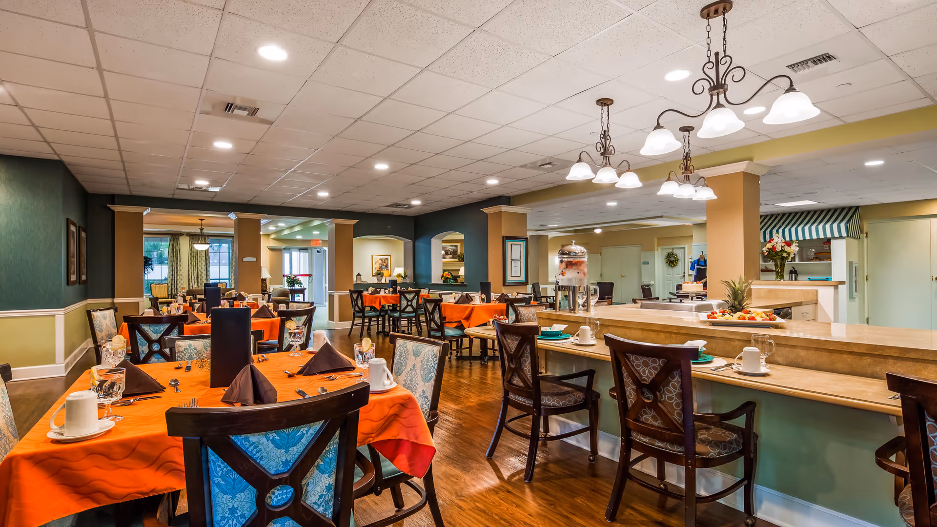 A spacious dining room with multiple tables covered in orange tablecloths, set with cups, glasses, and napkins. The room features wooden chairs with patterned cushions, a counter with additional seating, and warm lighting from ceiling fixtures. The walls are painted in green and beige tones, and there are decorative elements such as framed pictures and plants.