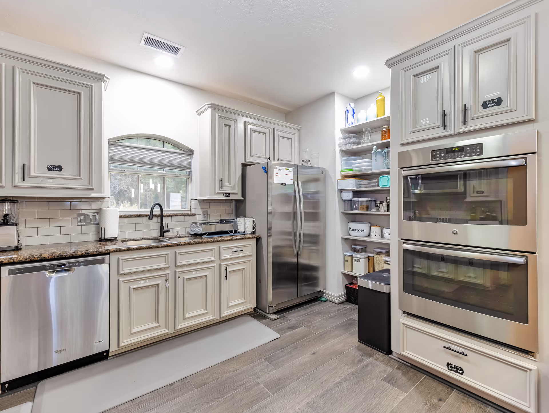 Modern kitchen with white cabinetry, stainless steel appliances including a dishwasher, double wall ovens, and a refrigerator. There is a window above the sink with a black faucet, granite countertops, and open shelving stocked with containers and kitchen items. The floor is wood-style and the space is well-lit with recessed lighting.