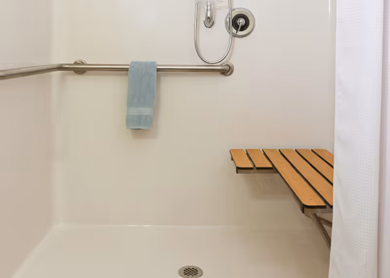 Accessible shower area with a metal grab bar, a light blue towel hanging on the bar, a wall-mounted showerhead, and a wooden fold-down shower seat. A white shower curtain is partially visible on the right side.