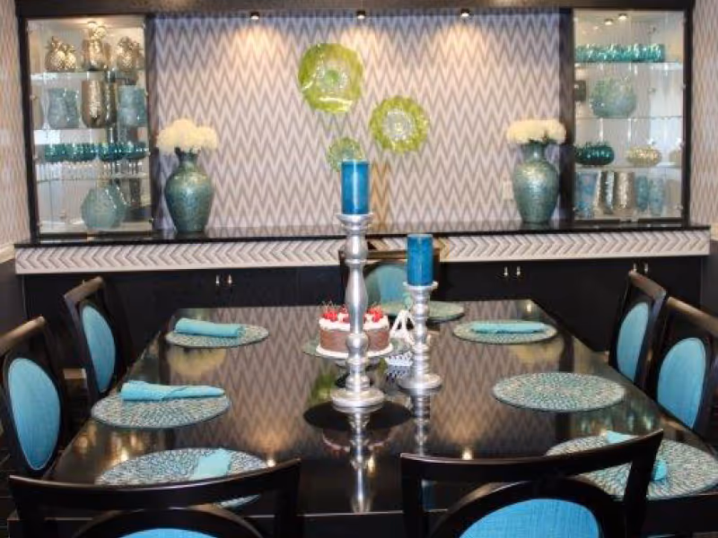 A dining room with a large rectangular black table set with turquoise placemats and matching napkins on black chairs with turquoise cushions. The table has three silver candle holders with blue candles and a small cake in the center. The back wall features a chevron patterned wallpaper with three decorative green glass plates and two large turquoise vases with white flowers on a black cabinet with glass shelves displaying various glassware and decorative items.
