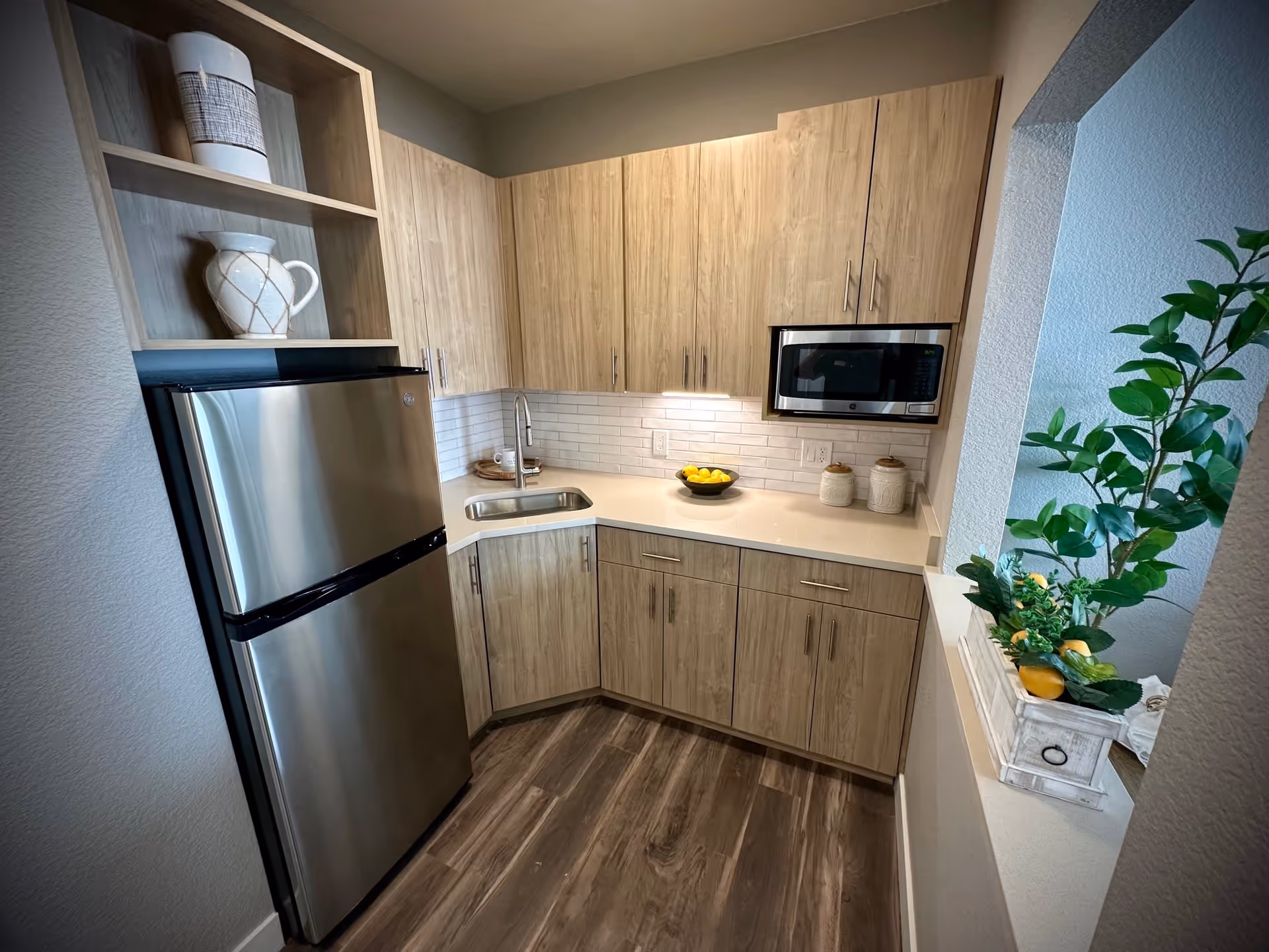 Small modern kitchen with light wood cabinets, a stainless steel refrigerator, a built-in microwave, a sink, and a countertop decorated with a bowl of lemons and two ceramic jars. There is a plant with green leaves and yellow lemons on the right side near an open wall space.