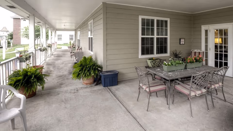 Covered outdoor patio area with a metal table and six cushioned chairs, decorated with flower boxes. Several potted plants and ferns line the concrete floor along the railing. A white plastic chair is visible in the foreground. The patio is attached to a building with beige siding and white-framed windows and doors.