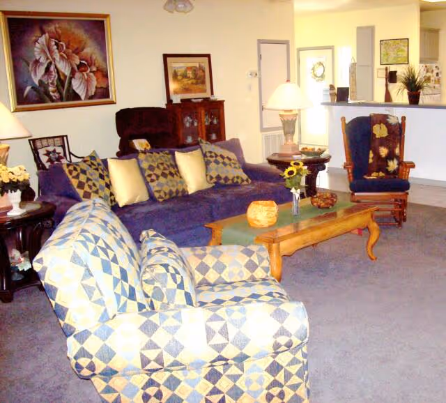 A cozy living room with a blue patterned armchair in the foreground, a blue sofa with multiple cushions, a wooden coffee table with decorative items, a wooden rocking chair with a cushion, and a side table with a lamp. The background shows a partial view of a kitchen area and a doorway.