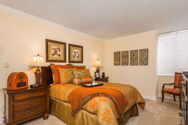 A warmly decorated bedroom with a bed featuring orange and gold bedding, two bedside tables with lamps, framed artwork on the walls, a window with blinds, and a wooden chair in the corner.