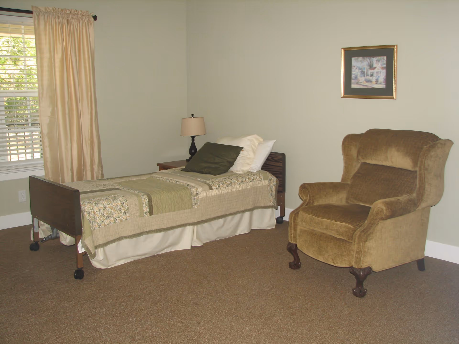 A simple bedroom with a single bed covered with a green and beige quilt, a wooden nightstand with a lamp, a large brown upholstered armchair, a window with beige curtains, and a framed picture on the wall.