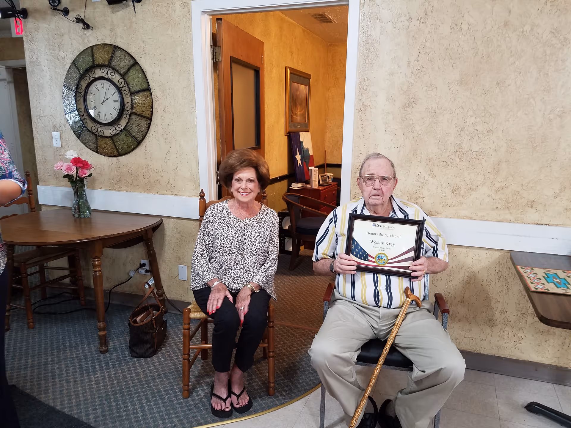 An elderly man and a woman sitting indoors in a room with beige textured walls. The man is holding a framed certificate and has a wooden cane resting on his lap. The woman is sitting beside him smiling. There is a round wooden table with a vase of pink flowers and a large decorative wall clock behind them.