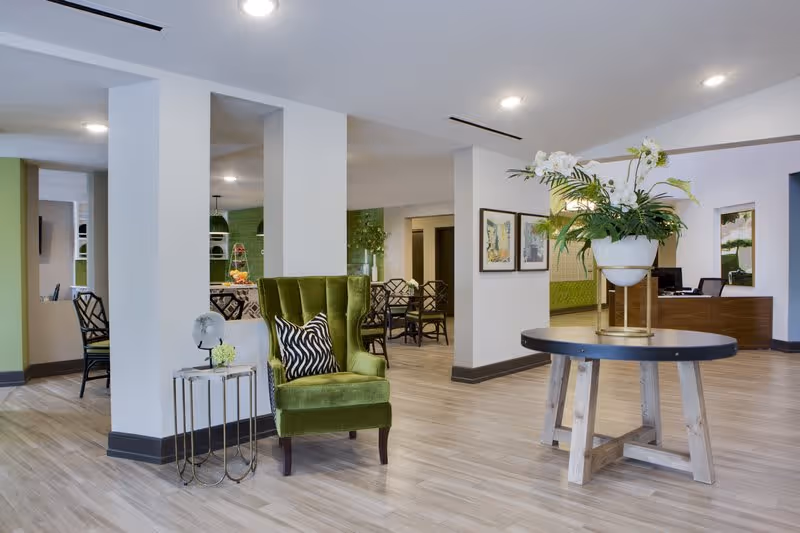 A spacious and modern interior area of Renaissance North Tampa featuring a green velvet armchair with a black and white patterned pillow, a small side table with decorative items, a round wooden table with a large white flower arrangement, and a reception desk in the background. The space has light wood flooring, white walls, and recessed ceiling lights.