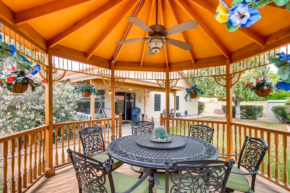 Wooden gazebo with a round metal table and chairs beneath a ceiling fan, surrounded by hanging flower baskets and a landscaped yard.