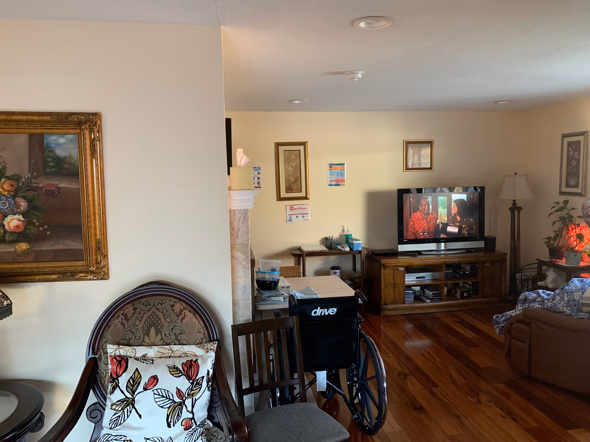 A cozy living room with wooden flooring, a TV on a wooden stand showing a scene with two people, a brown armchair with a blue patterned blanket, a wooden chair, a wheelchair, a side table with a lamp and plants, and framed artwork on the walls.
