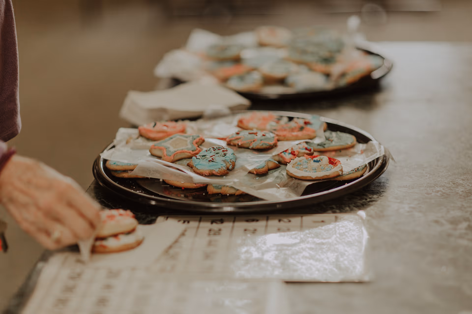 Close-up of decorated sugar cookies on a black tray lined with parchment paper, with a hand reaching for one cookie. The cookies are decorated with colorful icing and sprinkles in various shapes including stars and circles.