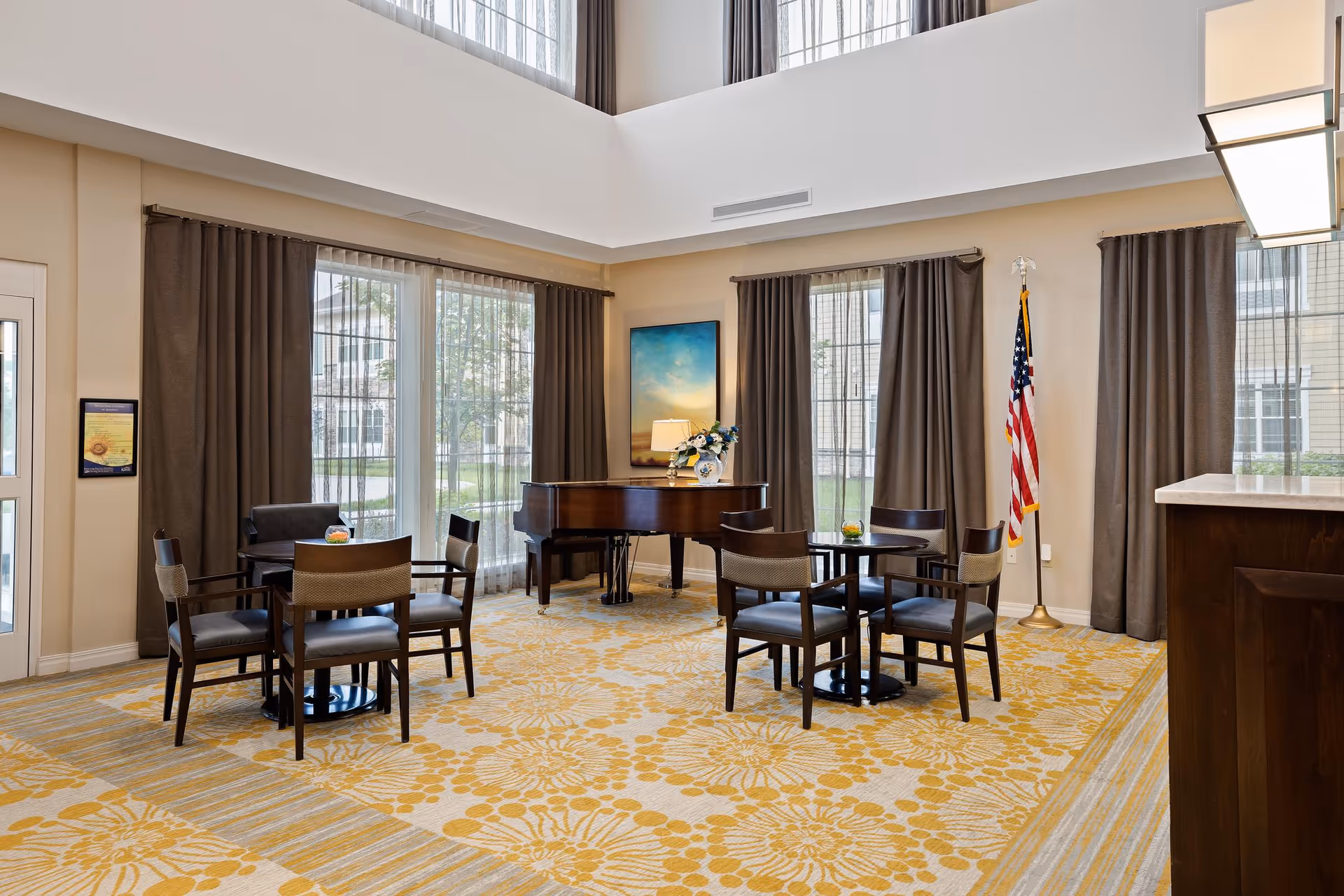 A bright and spacious common area with large windows covered by sheer and dark curtains. The room features a yellow floral patterned carpet, several small tables each surrounded by four chairs, a grand piano with a vase of flowers on top, an American flag in the corner, and a framed artwork on the wall.