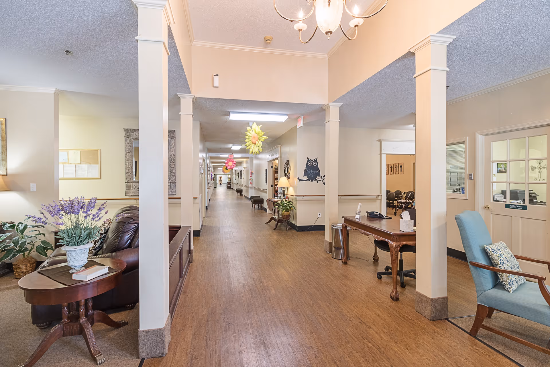 Bright interior hallway and seating area in a senior living facility with columns, desks, chairs, and decorative plants.