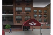 Exterior view of a multi-story brick building with a maroon canopy over the entrance displaying '300 CN&RC'. The building has several windows and a sign that reads 'Concord Nursing and Rehabilitation Center'. A person is walking on the sidewalk in front of the building.