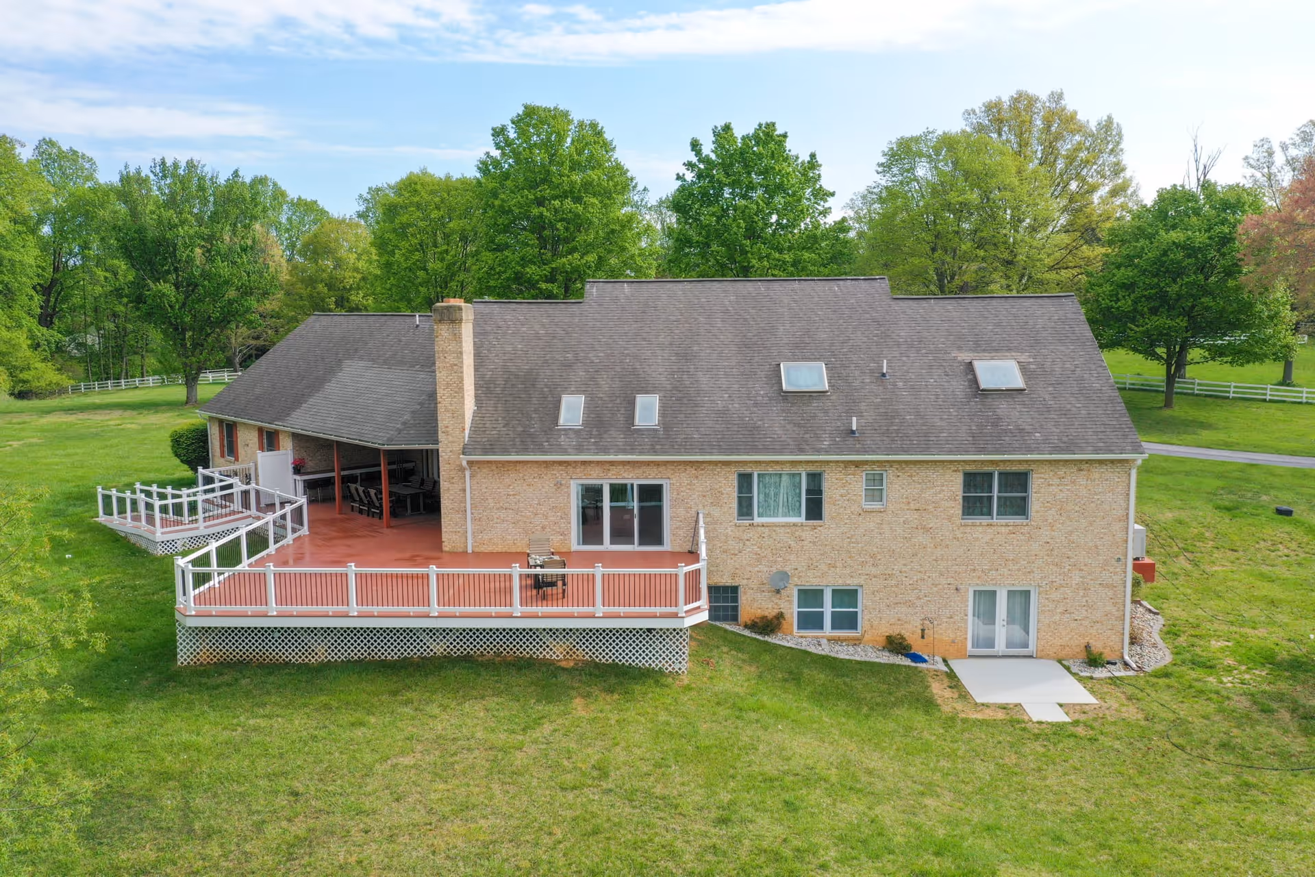 Rear exterior view of a large two-story brick house with a spacious red deck and white railing, surrounded by green grass and trees under a partly cloudy sky.