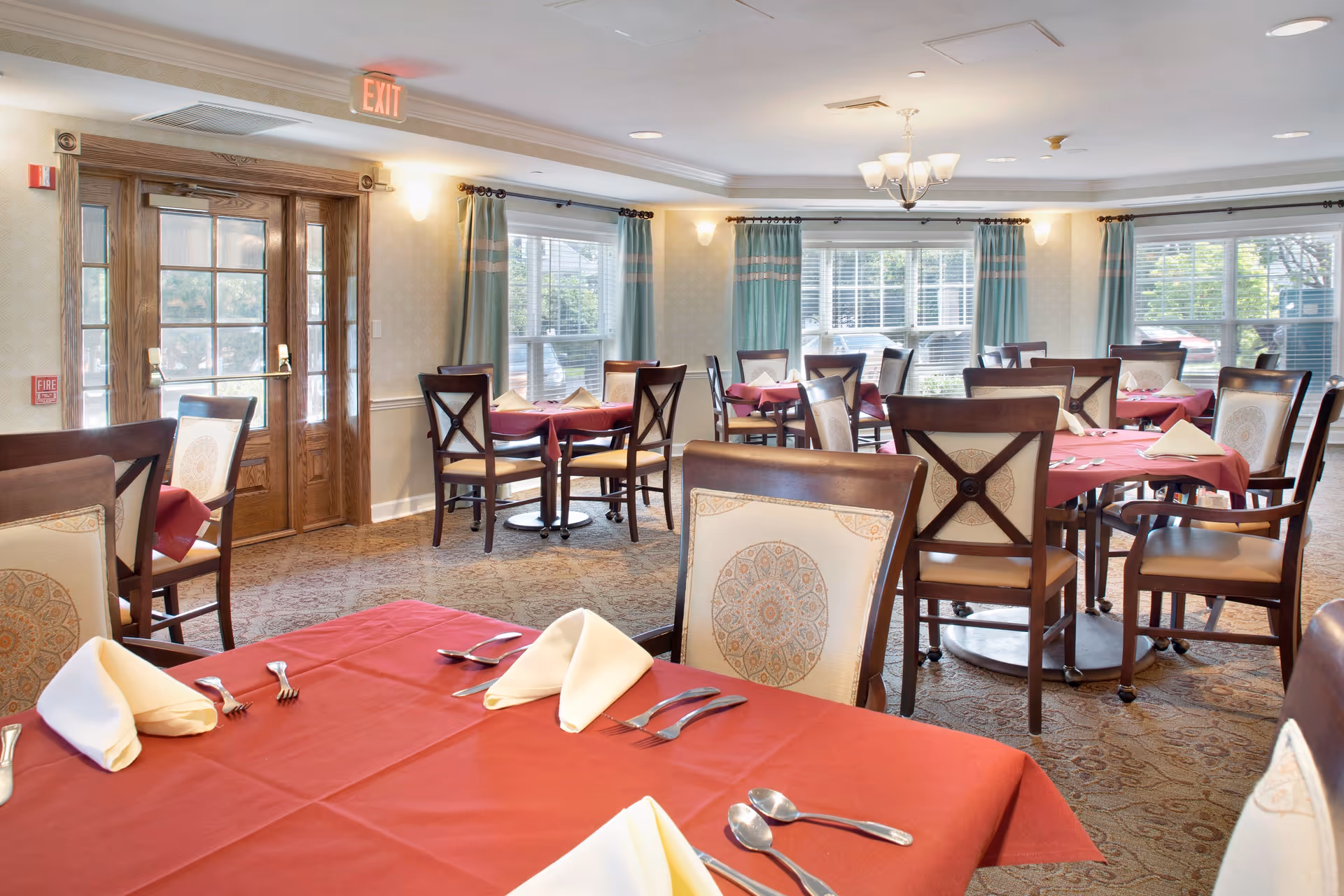 Bright dining room with tables set with red tablecloths and folded napkins, wooden chairs, large windows and double wooden doors.