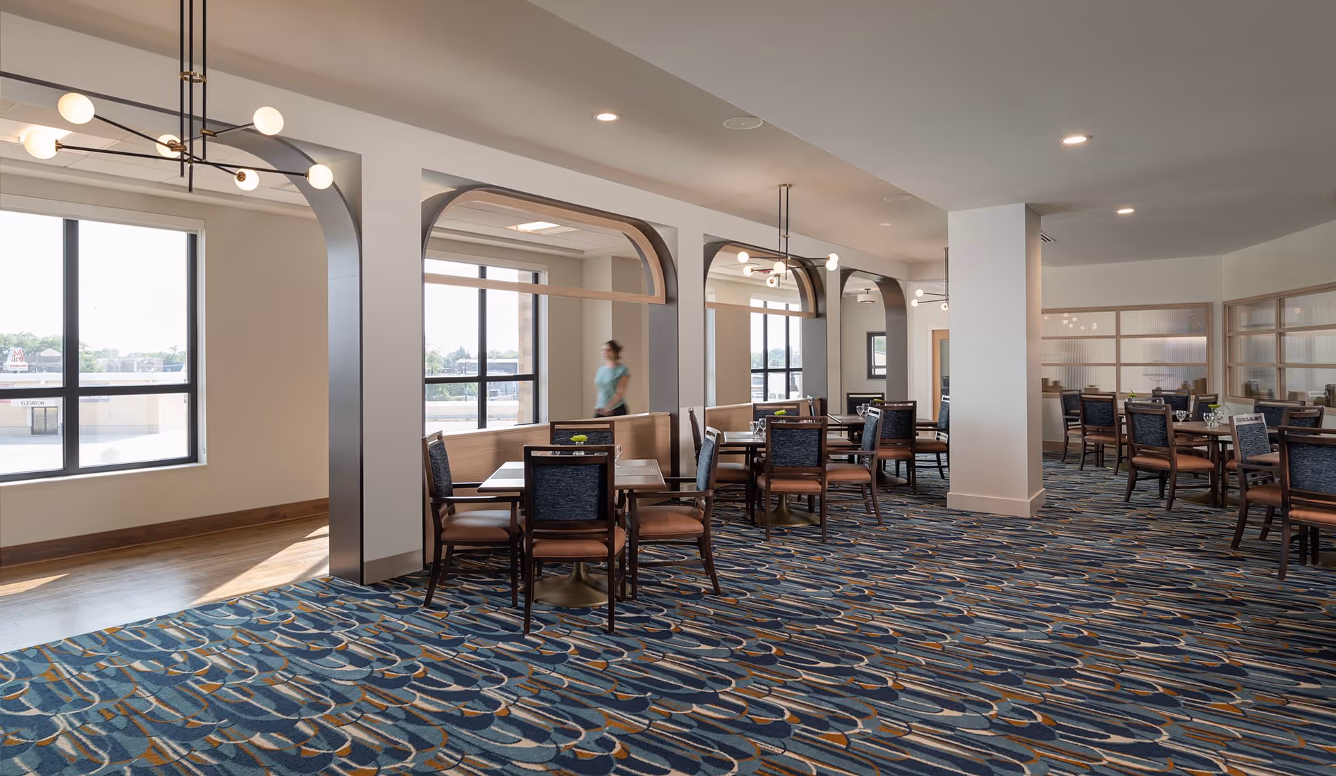 A spacious dining room with multiple tables and chairs arranged neatly. The room features large windows allowing natural light to fill the space, decorative arches, modern light fixtures, and a patterned carpet. A person is walking in the background near the windows.