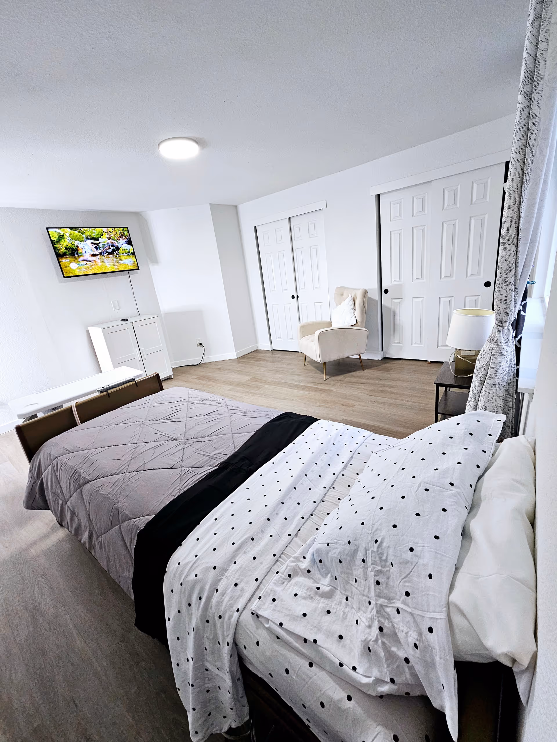 A bright and clean bedroom with a bed made with white and black polka dot bedding and a gray quilt. There is a beige armchair with a white pillow near two white sliding closet doors. A small black side table with a lamp is next to the bed. A flat-screen TV is mounted on the wall above a white cabinet. Light wood flooring and white walls complete the room.