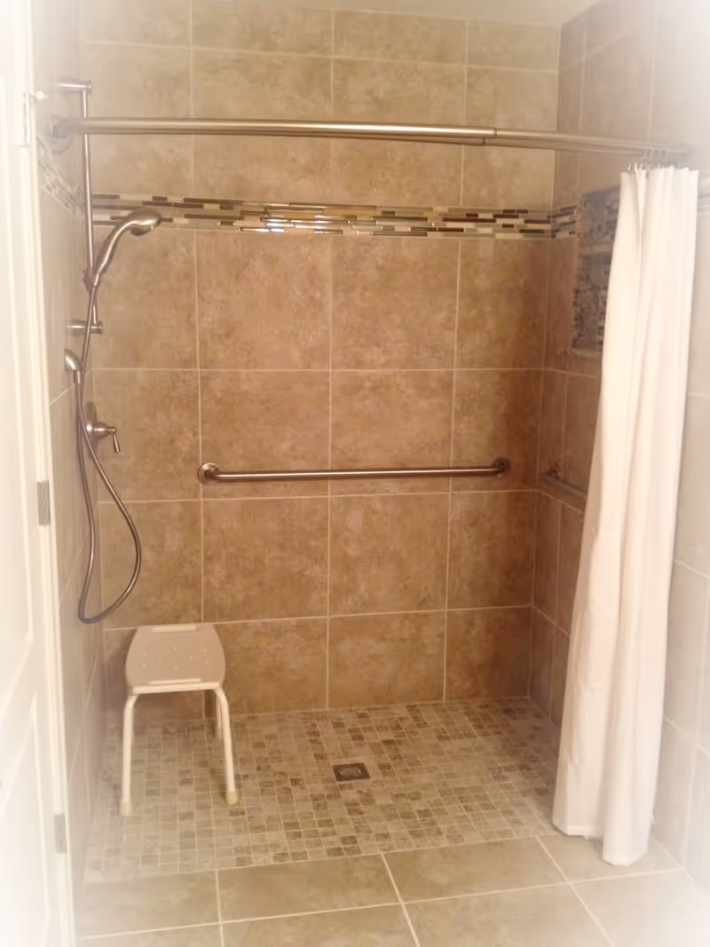 A tiled walk-in shower with a handheld showerhead, a horizontal grab bar, a white shower curtain, and a small white shower stool. The shower walls have a decorative horizontal tile accent strip.