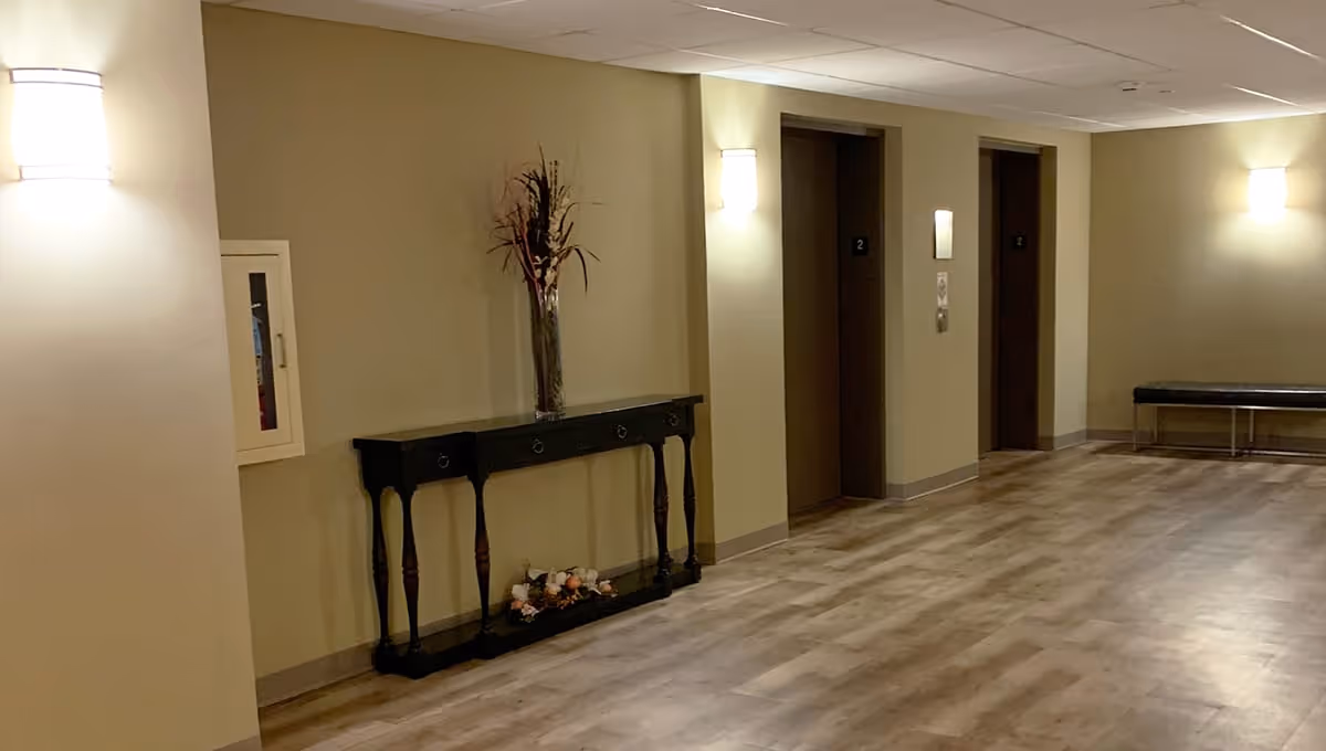 Interior hallway of Poplar Creek Village featuring two elevator doors, a black console table with a tall vase of dried flowers, wall-mounted lights, a fire extinguisher cabinet, and a black bench along the wall. The floor has a wood-like finish and the walls are painted beige.
