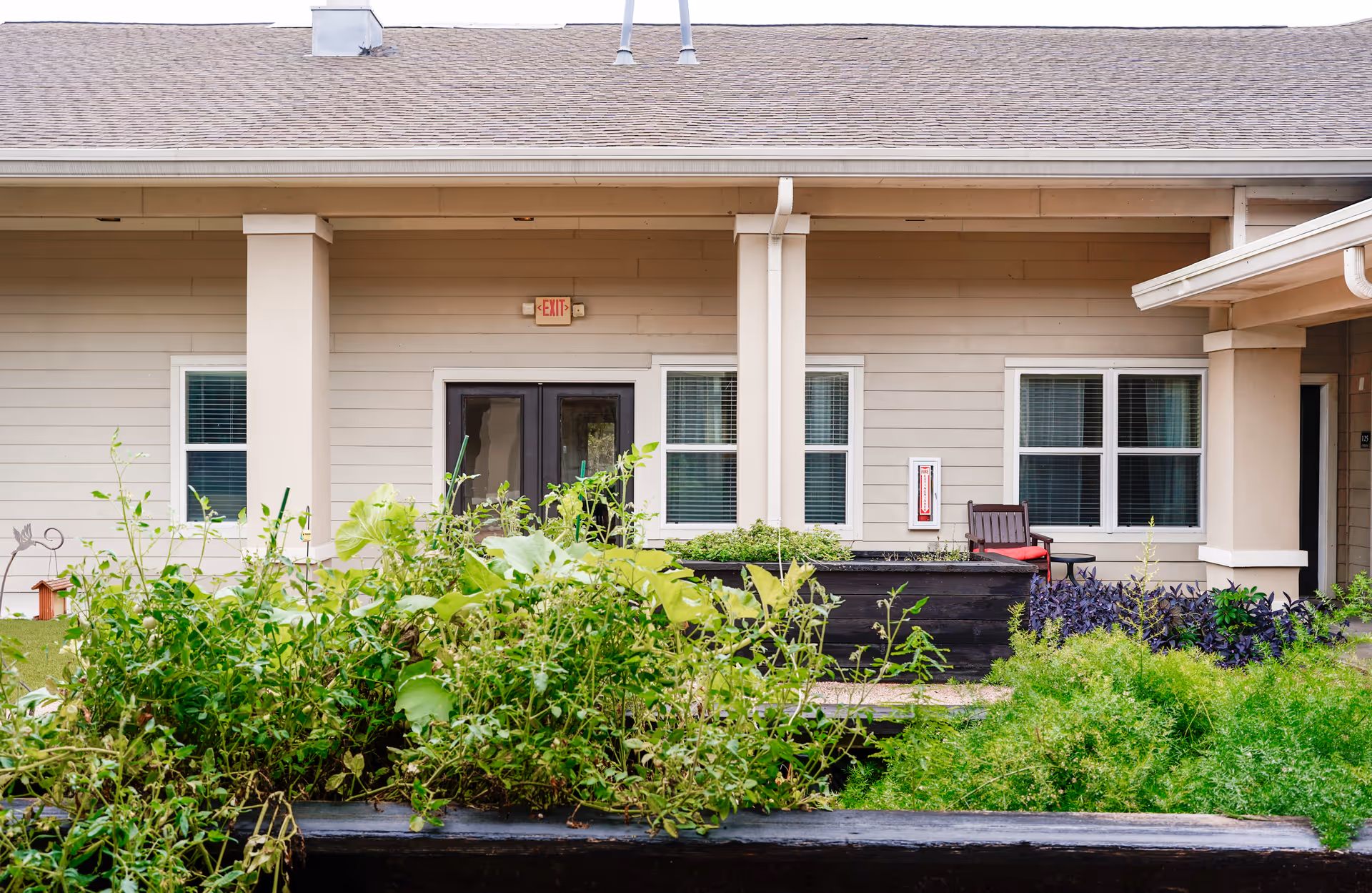 Exterior view of a beige building with a covered porch area, two windows with white blinds, a double door with an exit sign above it, and a garden with various green plants and shrubs in the foreground. There is a single chair with a red cushion near the windows.