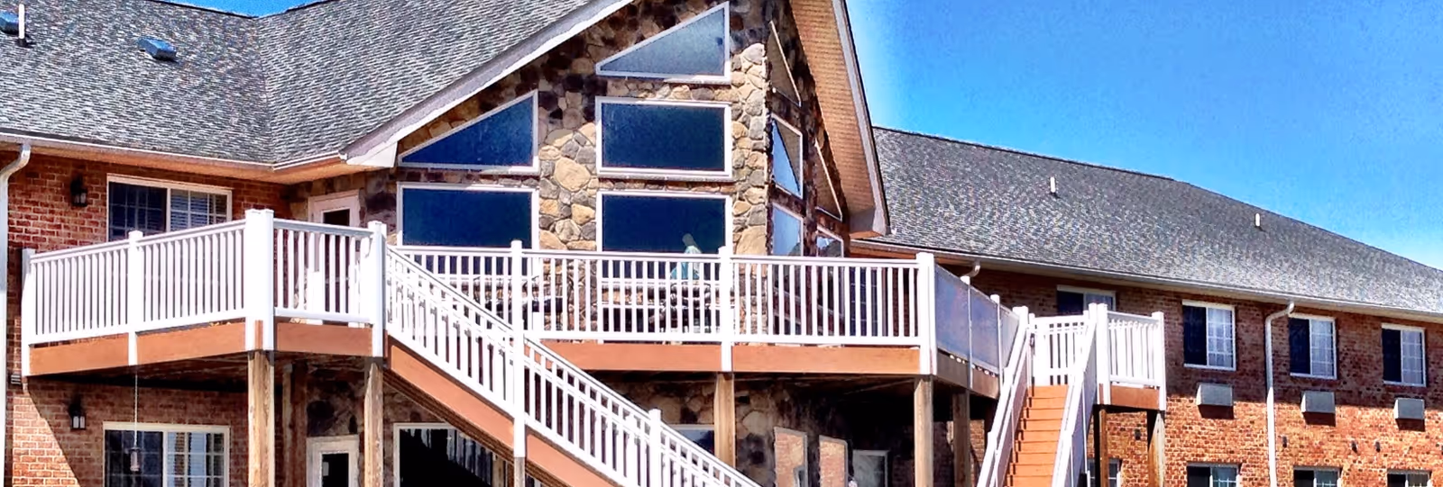 Exterior view of a brick and stone senior living building with large triangular windows, an elevated deck and staircases under a clear blue sky.
