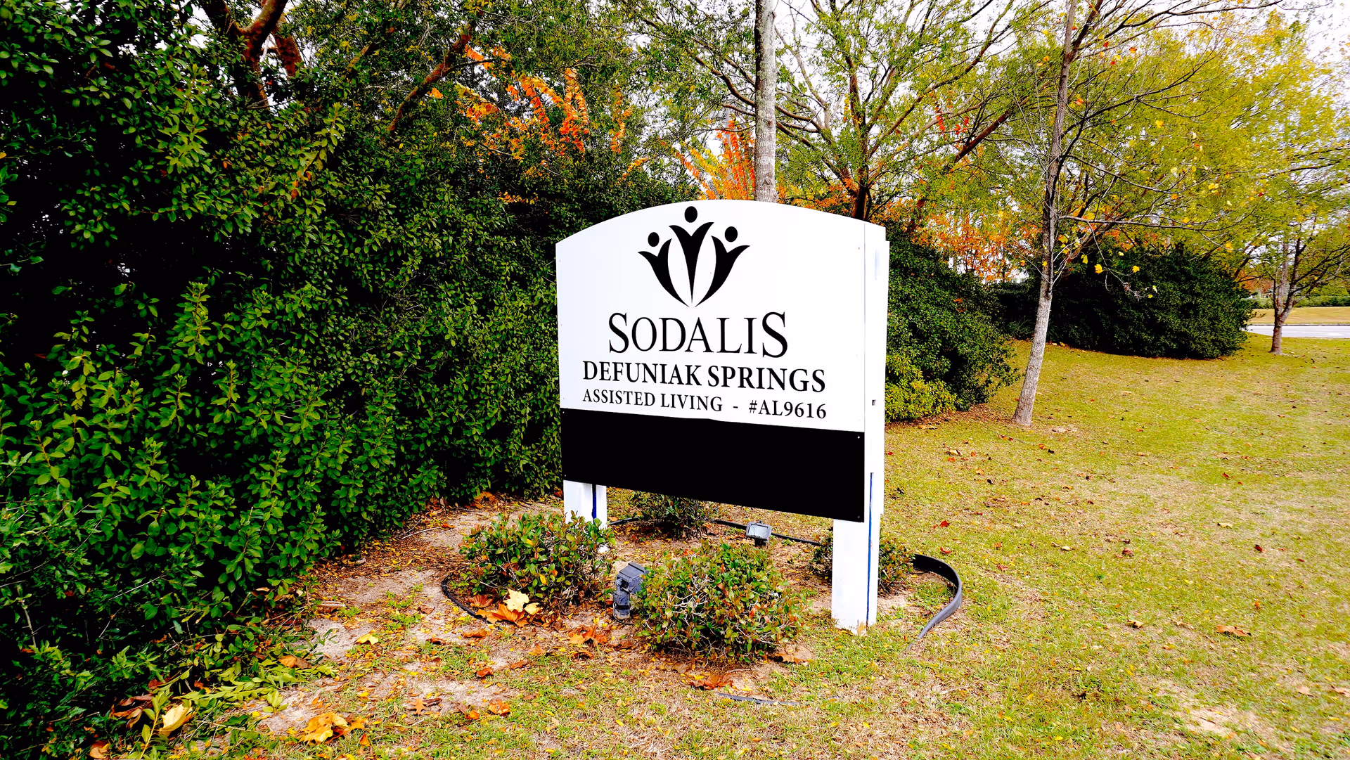 A white sign for Sodalis Defuniak Springs Assisted Living is placed on a grassy area with bushes and trees in the background. The sign features a black logo of three abstract human figures and text indicating the facility's name and assisted living license number.