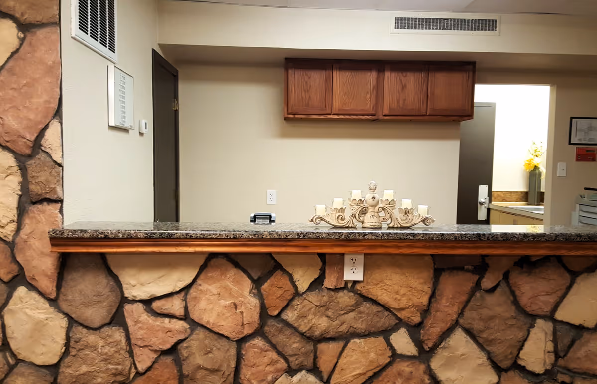 Reception desk area with a stone facade and a granite countertop. Behind the desk, there are wooden cabinets mounted on the wall, a door, and a small open window to another room with a vase of yellow flowers. A decorative candle holder with multiple candles is placed on the countertop.