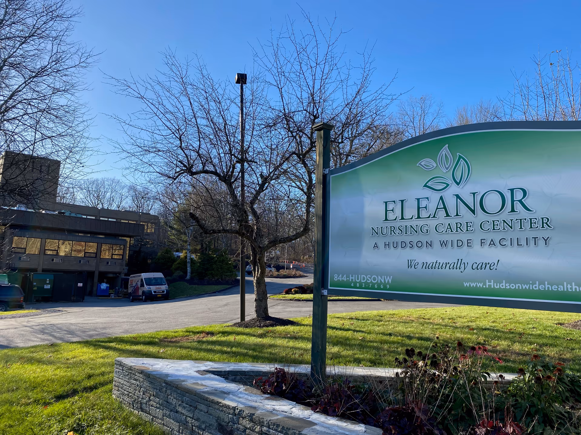 Outdoor view of the Eleanor Nursing Care Center sign with a building and leafless trees in the background under a clear blue sky.