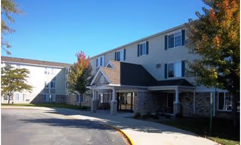 Exterior view of a two-story residential building with beige siding and multiple windows, surrounded by trees with autumn foliage under a clear blue sky. The building has a covered entrance with a peaked roof and a driveway in front.