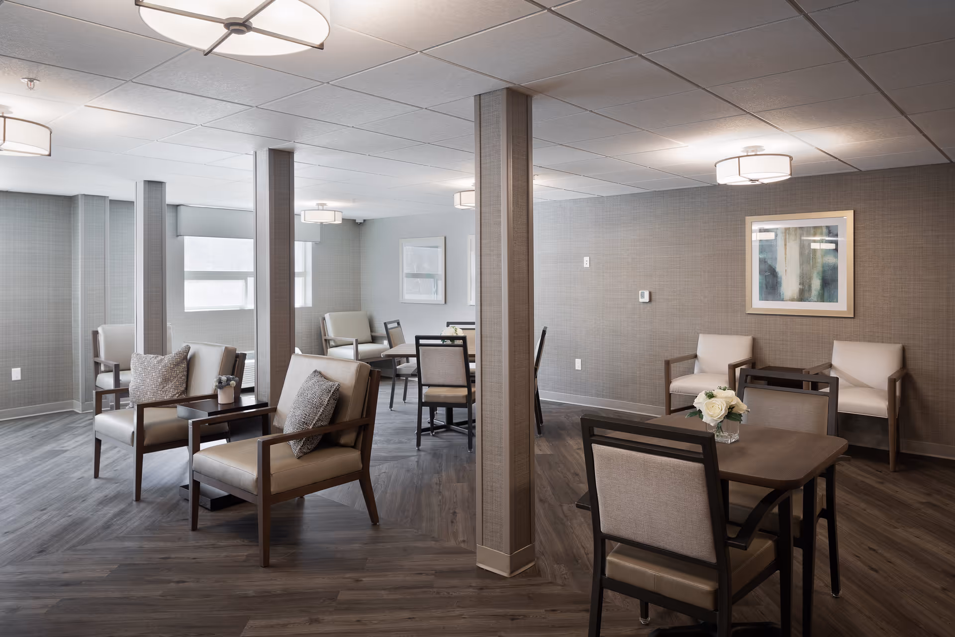 Spacious, well-lit communal lounge with upholstered armchairs, dining tables, and wall art.