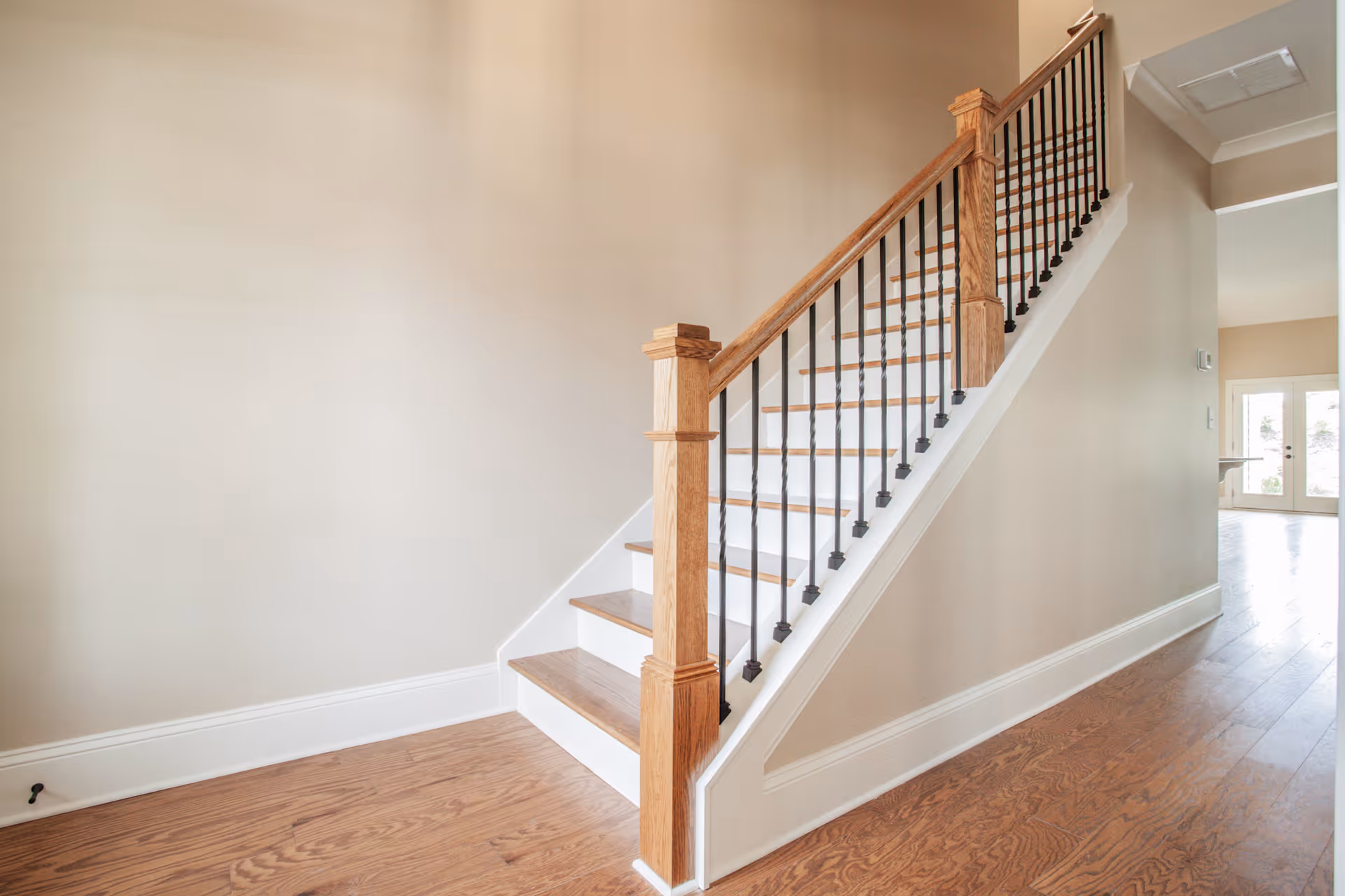 Interior view of a staircase with wooden steps and black metal balusters, adjacent to a beige wall and hardwood floor, leading to an upper floor with a hallway visible in the background.