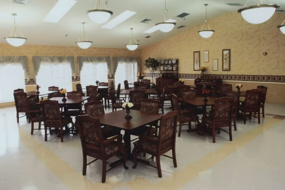 A spacious dining room with multiple dark wooden tables and matching chairs arranged neatly. Each table has a small floral centerpiece. The room has large windows with decorative curtains allowing natural light to fill the space. The walls are adorned with framed artwork and a decorative border. Ceiling lights hang evenly spaced across the room.