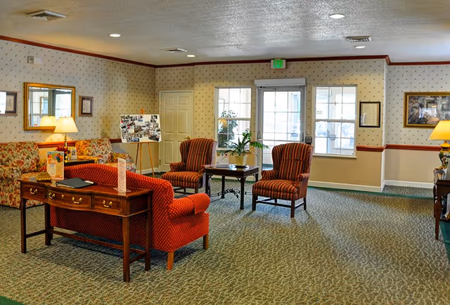 Spacious senior living facility common room with upholstered armchairs and sofas arranged around tables near windows and exit doors.